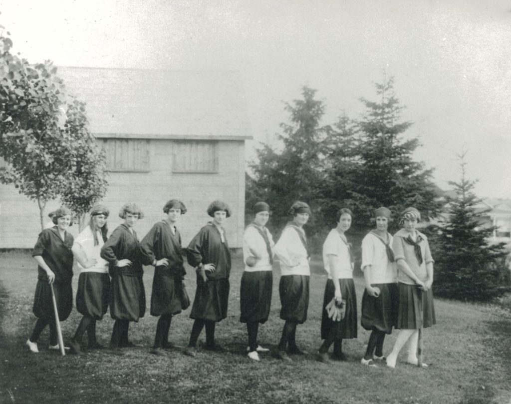 Ladies Softball Team at Alexandra Park, 1924 A lineup of young women pose in their softball uniforms. Some of the women have softball batt or mitt in their hands. Behind them is a wooden house and tall pine trees.