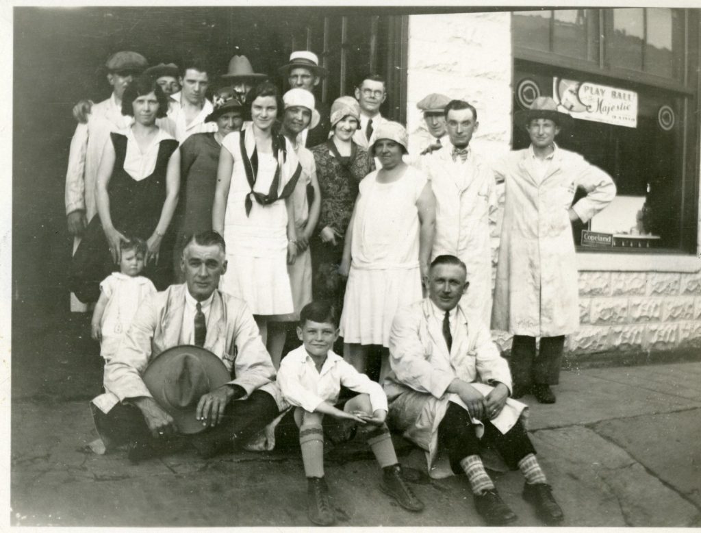 Group Gathered for Chapman's Auto Parade, c. 1929 A group of men, women, and children pose for a photo on the sidewalk outside a building. Most of the men are wearing long coats with either a tie or bowtie. The women wear dresses that reach their knees. There are two children in the image; a young boy sits on the ground in between two men, and the other is a toddler, standing yet being held by a young woman.