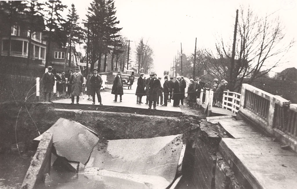Harmony Bridge Damaged by Flood, 1929 Set in townhouse lined street, group of people stretches across the edge of a collapsed bridge. The debris lies in the foreground and the area is roped off from the group.