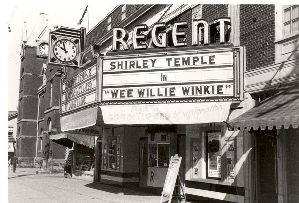 Regent Theatre, c. 1930 The front of the theatre is a part of a brick building lined street. In front, is the ticket booth, which has posters and signs lined up along the wall. The marquee above the theatre entrances reads, "Shirley Temple in 'Wee Willie Winkie'". Above the marquee, the word, "Regent" is written in capital letters and with lights. Below the marquee is a cloth sign reads, "Air Conditioned by Refridgeration".