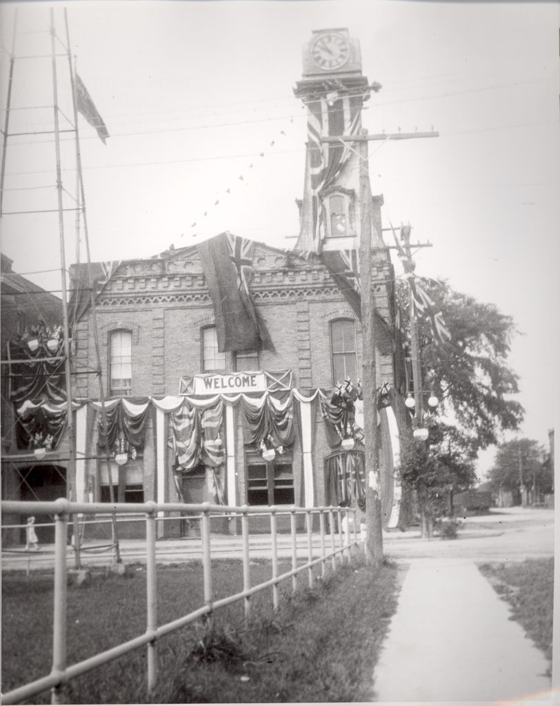 Town Hall, 1924 Across a short sidewalk, the side view of a two-story building with a clock tower is draped in bunting and flags. Underneath a second-story window, a sign reads, "Welcome".