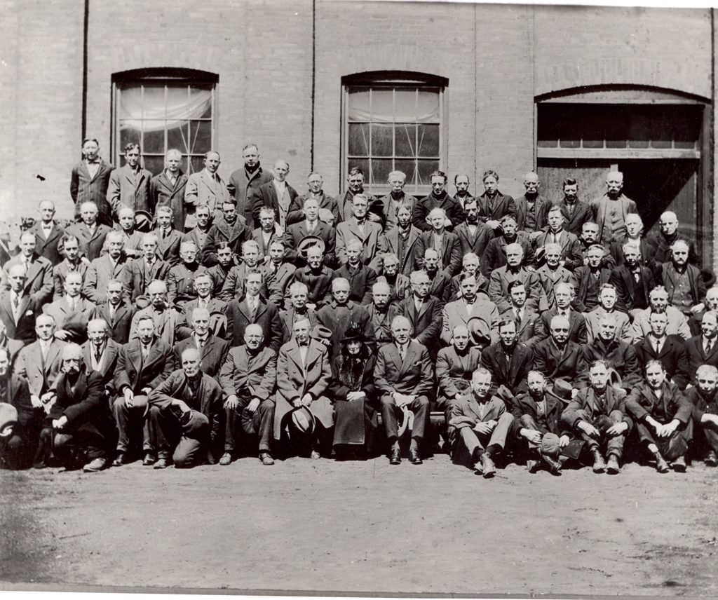 McLaughlin Employees of 20 Years’ Service, 1924 This large group portrait features men in 5 rows. They are all dressed in suits. Behind them is the factory building.