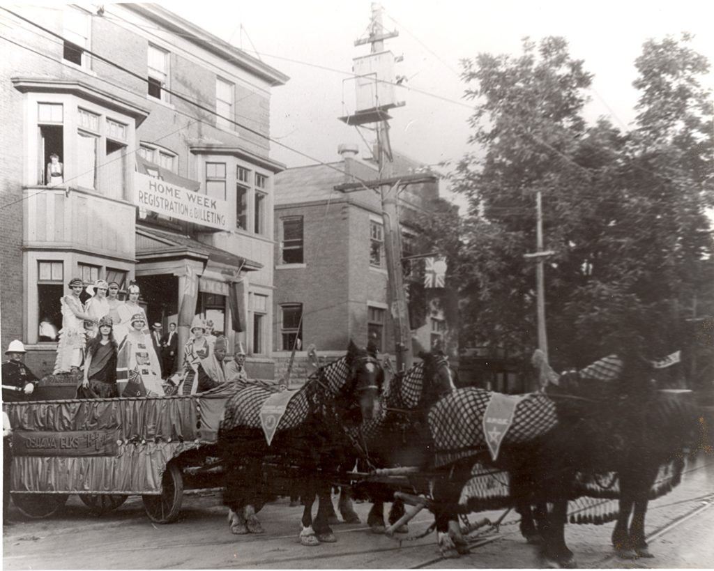 Old Home Week Parade, 1924 A float full of well dressed people is carried by four black horses. On the side of the of the float, a sign reads, "Oshawa Elks". Behind them is a three-story building with a banner running between two windows that reads, "Home Week Registration and Billeting".
