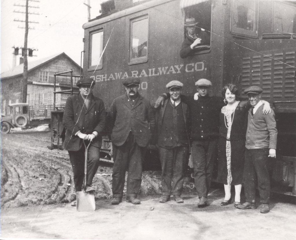 Railway Employees, 1920 Five men and one woman stand in front of a locomotive. The men are dressed in vests and ties, and the woman wears a dress that reached her ankles. The man to the far left has a shovel in his hands which he uses to perch up one foot. In the locomotive behind them, a man leans outside an open window. Along the locomotive reads, "Oshawa Railway Co.". In the background, a truck is parked in front of a wooden house.