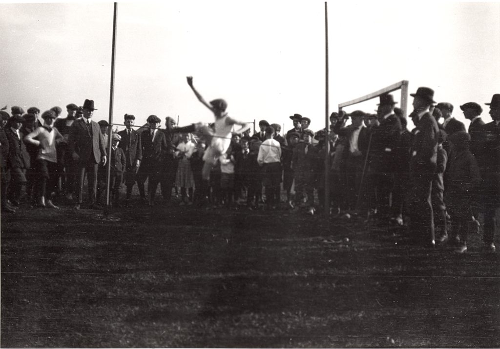 High Jump at a School Field Day, 1920 A group of people form a semi circle around the high jump pole. The high jump pole is made of two posts about eight feet apart and a pole running between the two. The group is mostly made up of men, adolescent to young adult, dressed in suits, coats and hats. The student jumping the pole is a adolescent boy, dressed in a tank top and shorts. He is a blur as he jumps the pole with his left leg first.