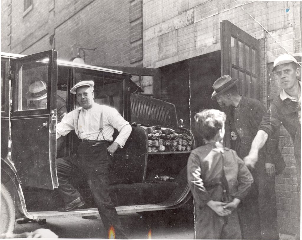 Rum Running During Prohibition, 1920 A man in suspenders stand with one foot in the car door. He has his hand on his hip and looks directly at the camera. Behind the car is an open garage and we can see the cargo of rum bottles in the back of the car. In the foreground, men peer into the car and garage. A boy stands with his back to the camera; to the right an young man attempts to pull him out of the way while looking at the camera.
