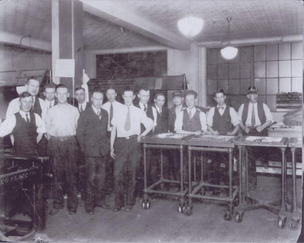Oshawa Daily Times Night Staff, 1928 A group of young men, dressed in suits and varying vests and ties, line up and pose for the photo. In front of the group are three wheeled trays that appear to work spaces, completed with papers and pens or pencils.