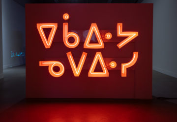 Orange neon channel sign hanging on a gallery wall that reads ekawiya nepewisi (don’t be shy).
