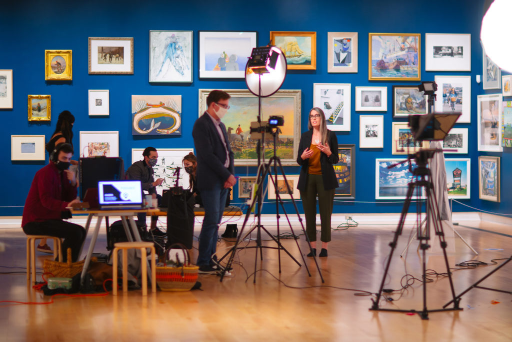 A group of people filming a video in front of a blue wall with many artworks hung on it.