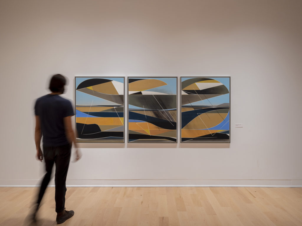 A person standing in front of three paintings on a white gallery wall.
