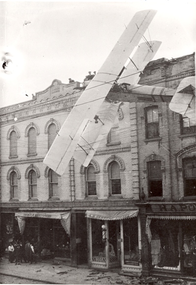 A black and white photograph of a small airplane flying in front of a building.