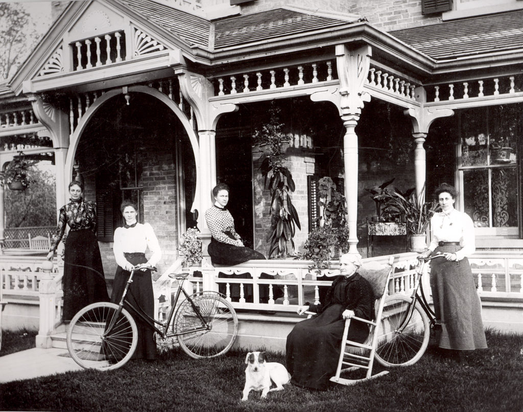 A black and white photograph of 5 women standing in front of a house with a white porch. There is a small dog sitting beside a woman in a rocking chair. 
