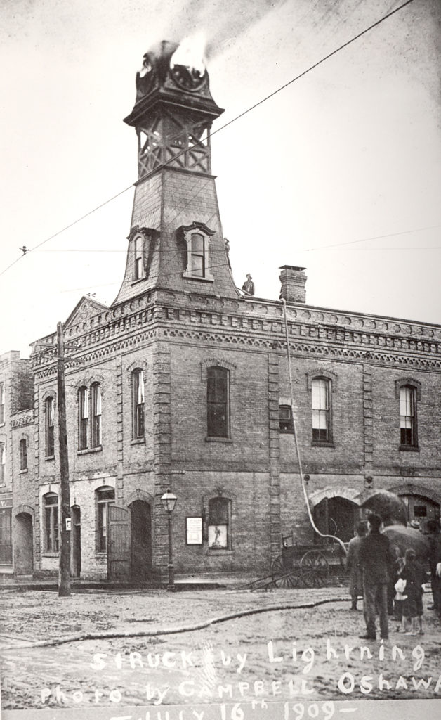 A black and white photograph of the town hall building with a tower on top of it while people walk down the street in the foreground. 