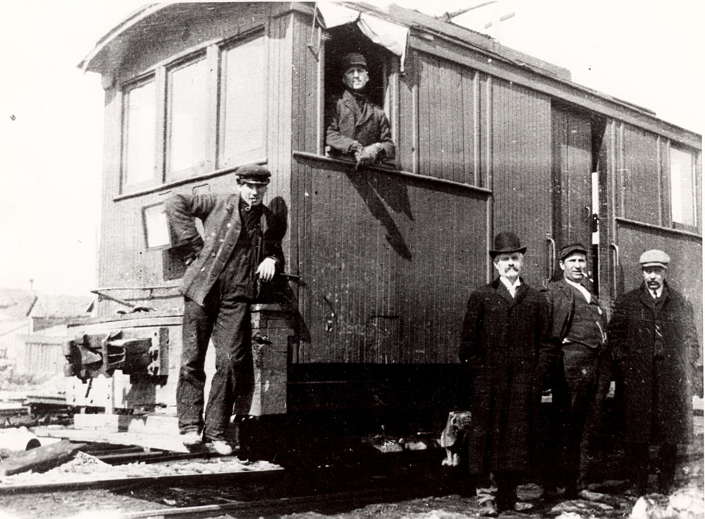 A black and white photograph of 5 men wearing suits standing around a train on the train tracks.