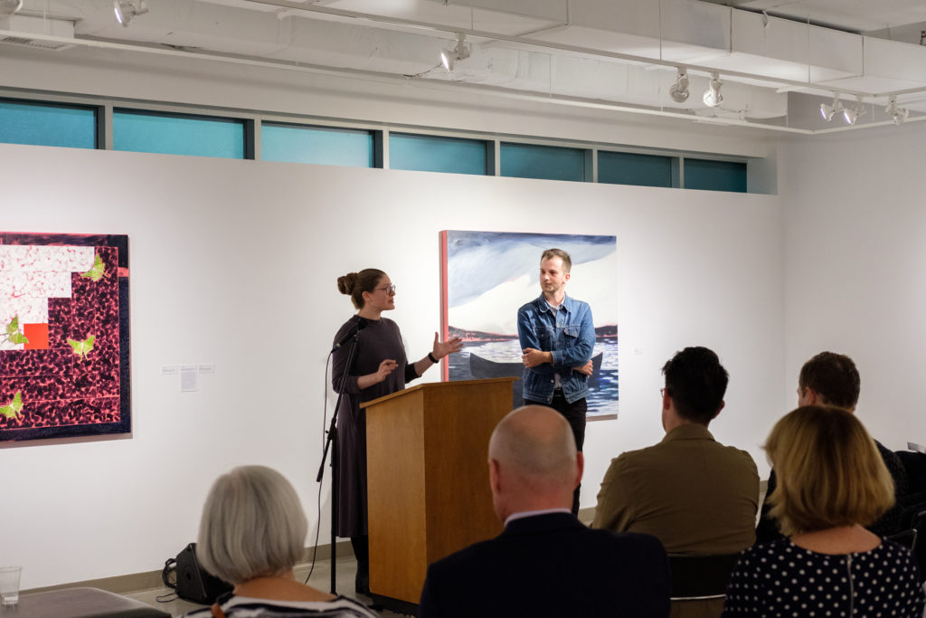 Two people in front of a podium presenting to a crowd with artworks hung on the wall behind them. 